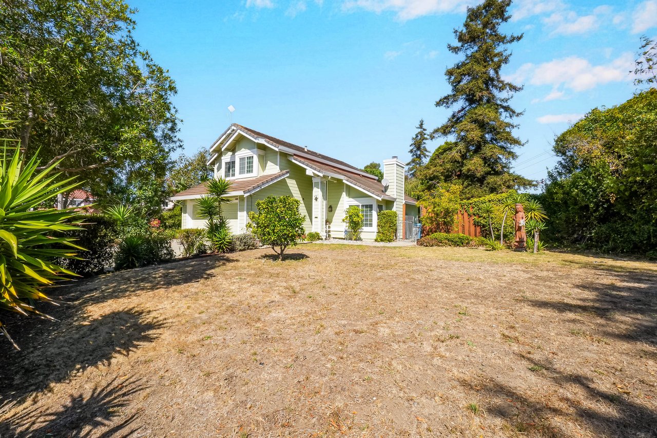 Light-colored two-story house beyond a broad dry lawn, framed by mature trees and dense landscaping.