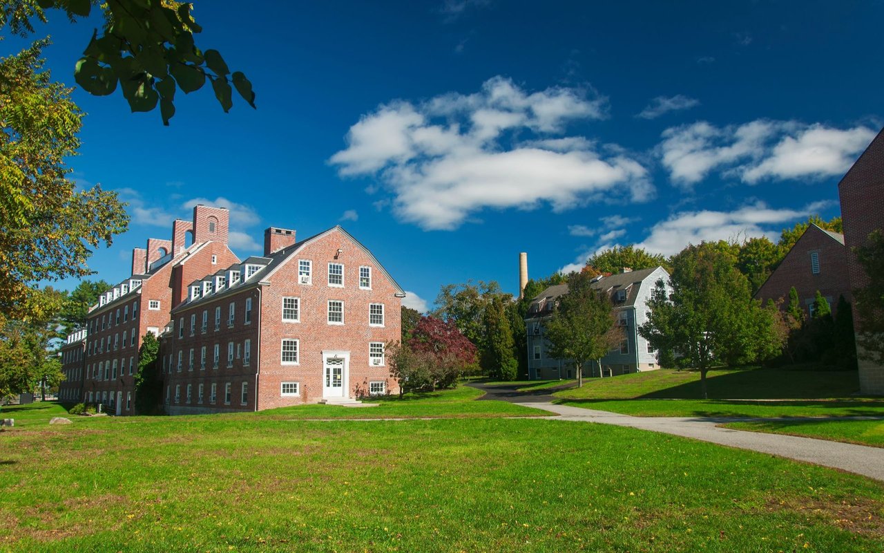 Campus-like brick buildings beside a broad lawn under a blue, cloud-dotted sky.