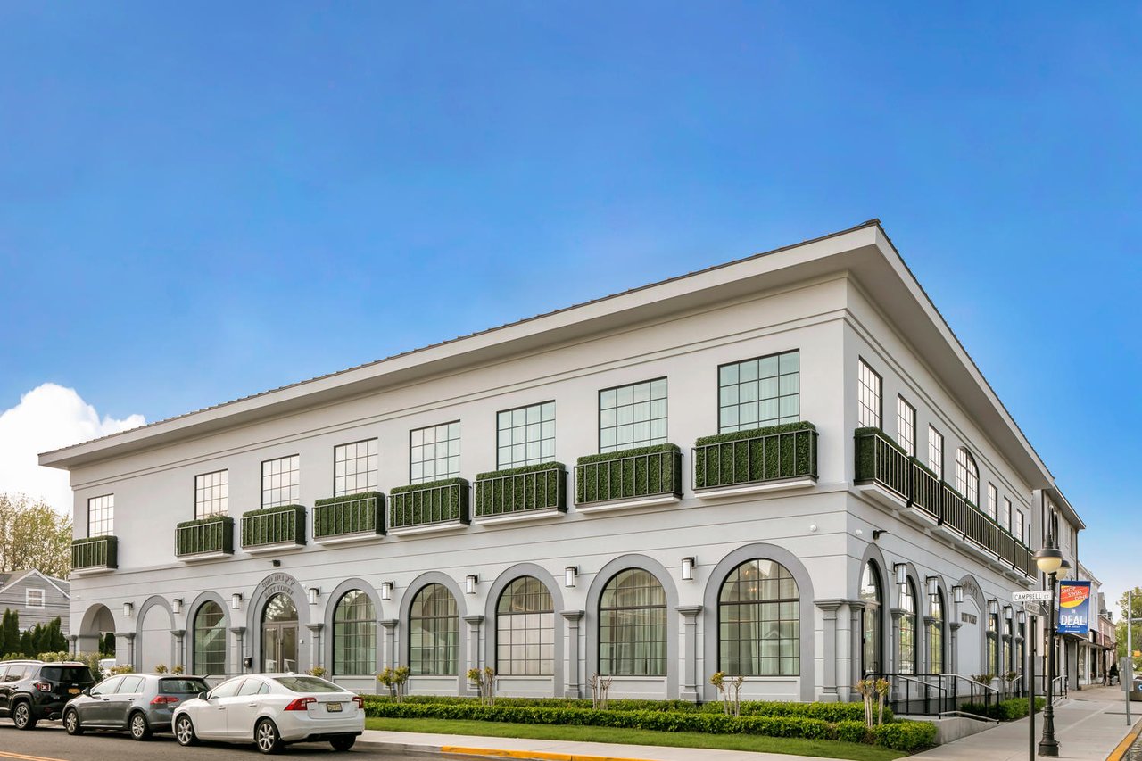 White two-story commercial building with arched windows and planted balconies along a street.