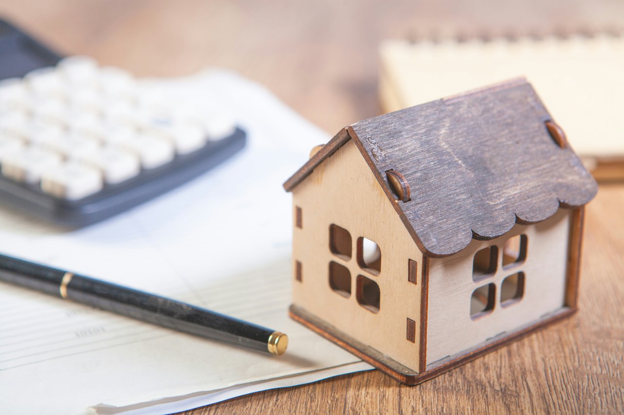A small wooden house model next to a calculator, pen, and financial documents on a desk