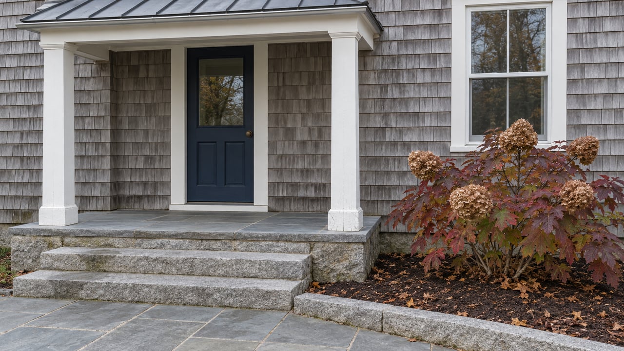 Weathered cedar-shingle home with a navy front door, white porch posts, granite steps and burgundy oakleaf hydrangea.