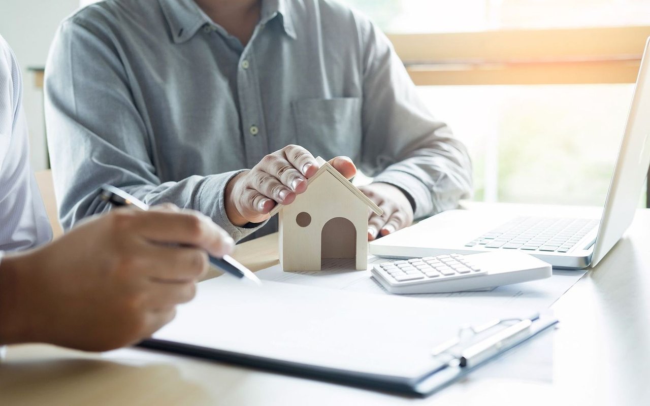 People discuss a home sale over paperwork, a calculator, and a small house model.