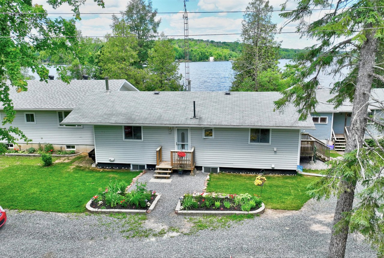 Gray-sided lakeside house with flower beds, gravel driveway, and water visible beyond the roof.