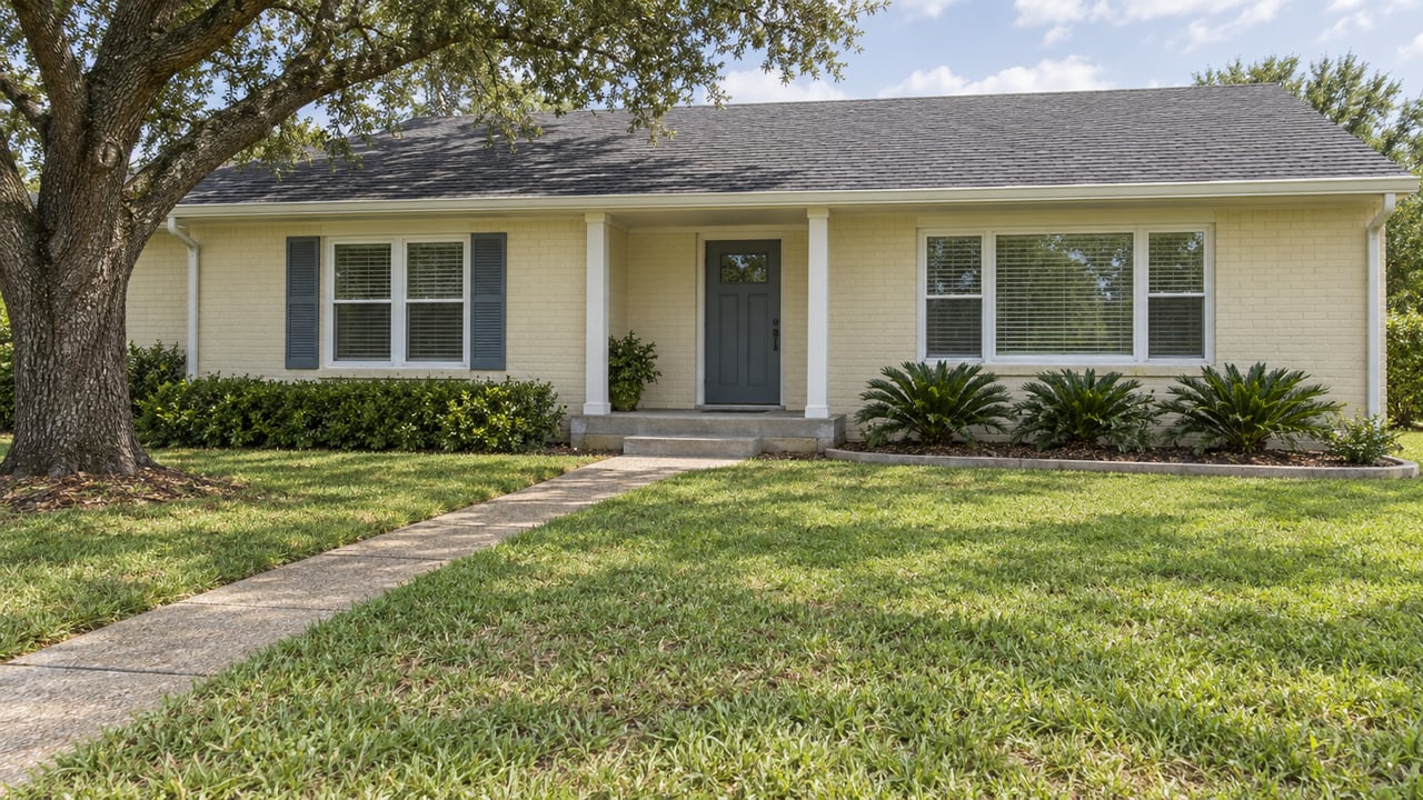 A pale painted-brick single-story house with a shallow front porch, blue-gray front door, deep lawn and mature oak.