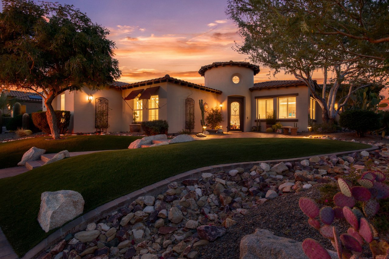 Front exterior of a stucco single-story home with desert landscaping and warm interior lights at sunset.