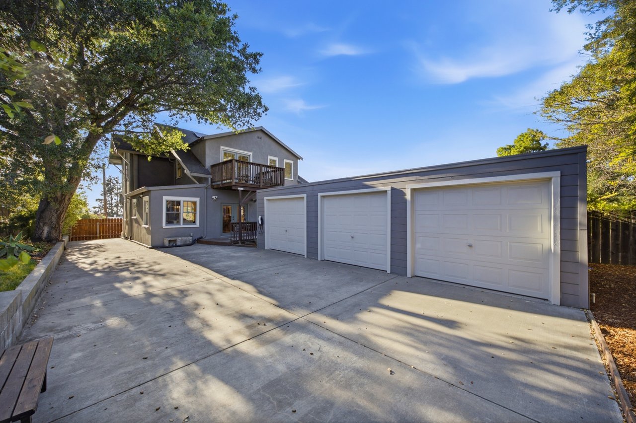 Gray two-story home with a balcony, three-car garage, and broad tree-shaded driveway.