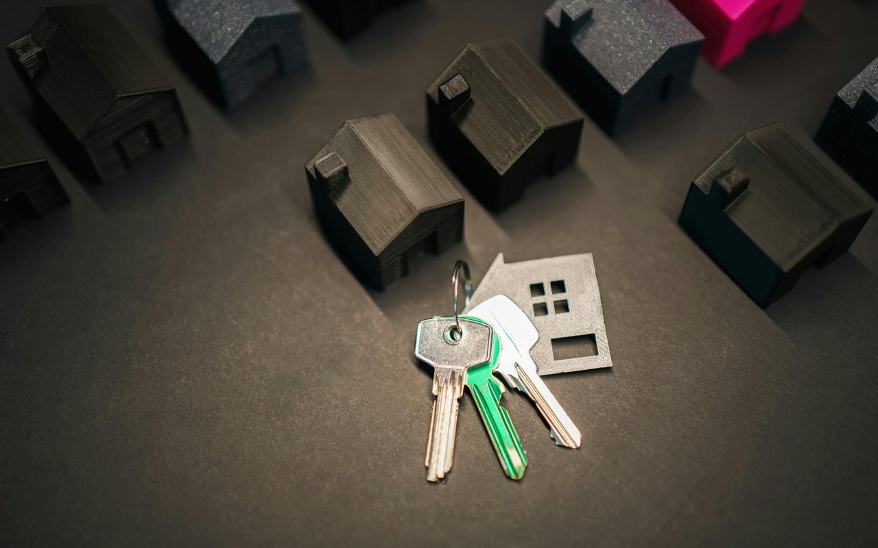 House keys beside a house-shaped cutout, with miniature homes in the background.