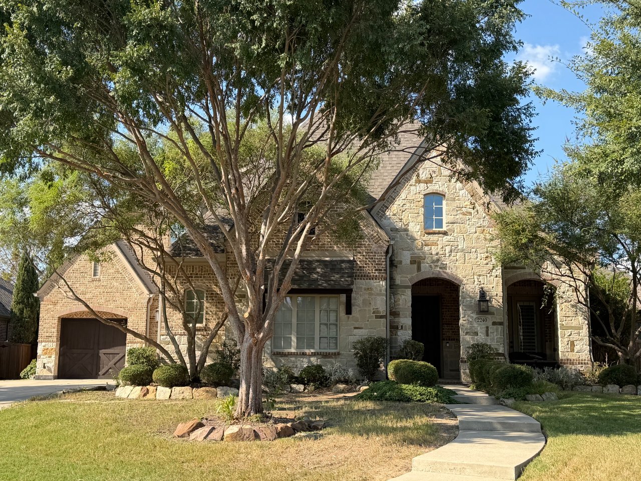 Two-story stone-and-brick house with a large front-yard tree and curved walkway.