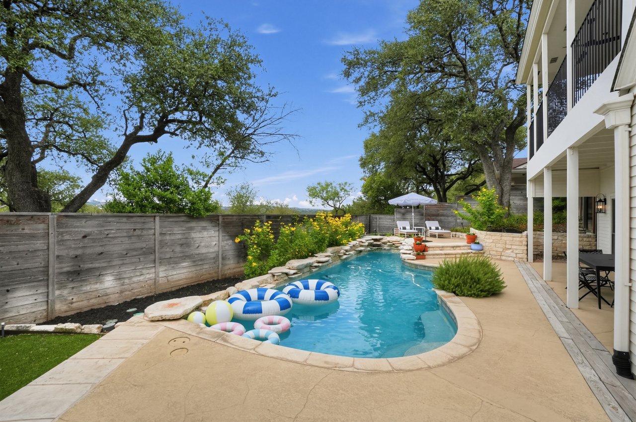 Backyard swimming pool with colorful floats, stone coping, and a patio beside a white house.