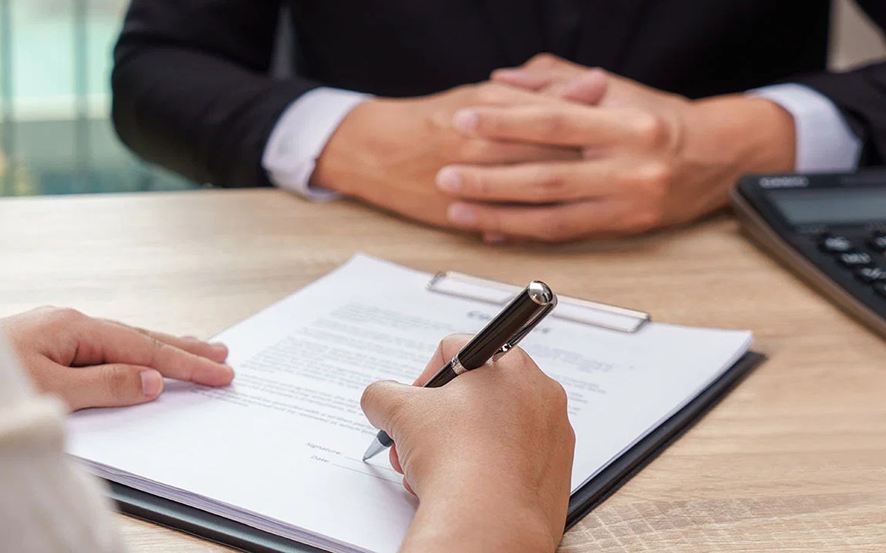 A person signs a document on a clipboard across a desk from someone with clasped hands.