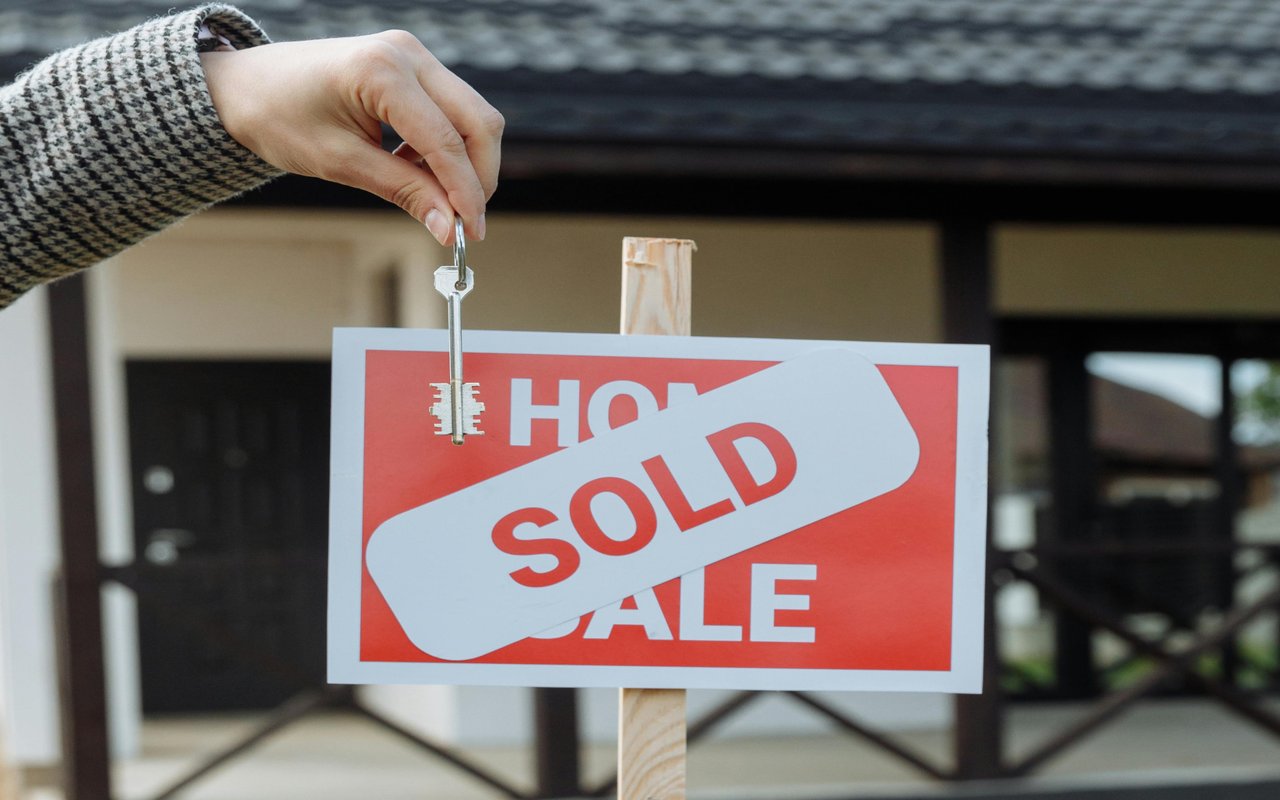 A SOLD sign in front of a house, with a hand holding a key above it.