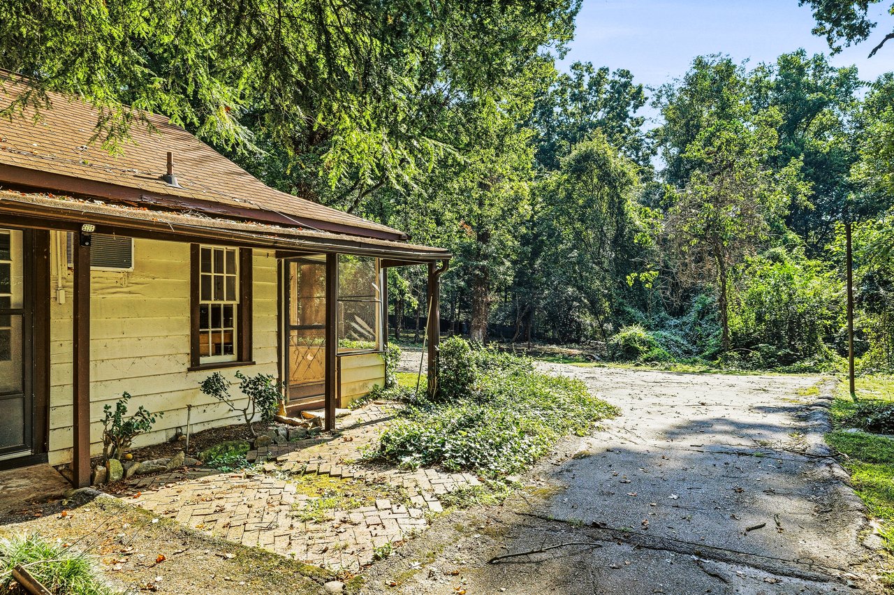 Single-story house with a covered porch beside a cracked driveway, surrounded by dense trees.