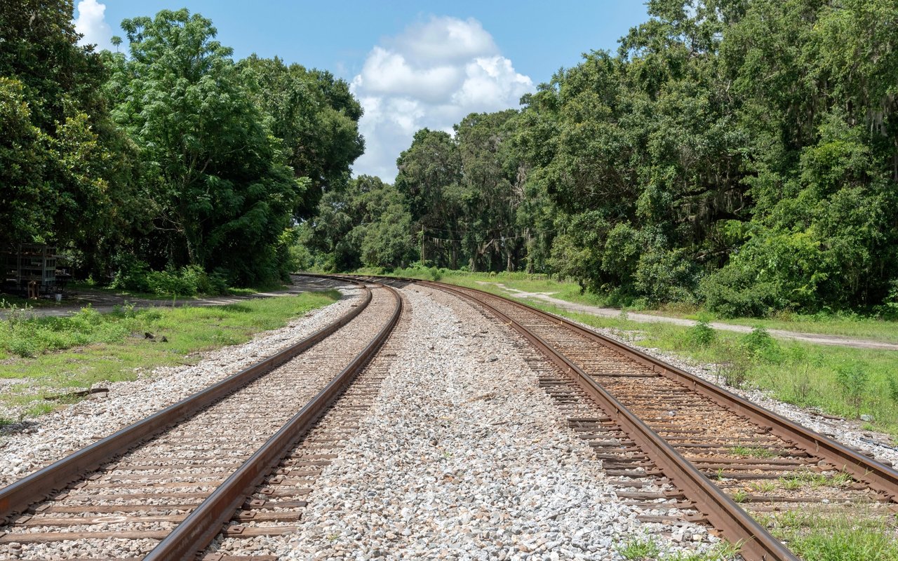 Railroad tracks curve through dense green trees beneath a partly cloudy sky.