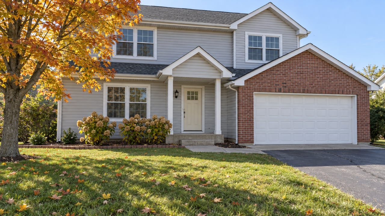 Two-story gray-sided home with red-brick garage accents, a deep autumn lawn, and an orange-yellow maple beside the entry.