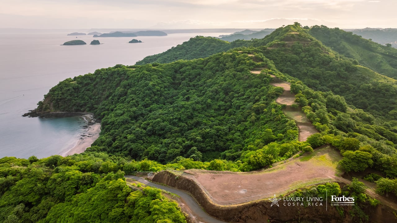 Coastal residential lot with cleared areas and access road amid forested hills overlooking the ocean and islands.