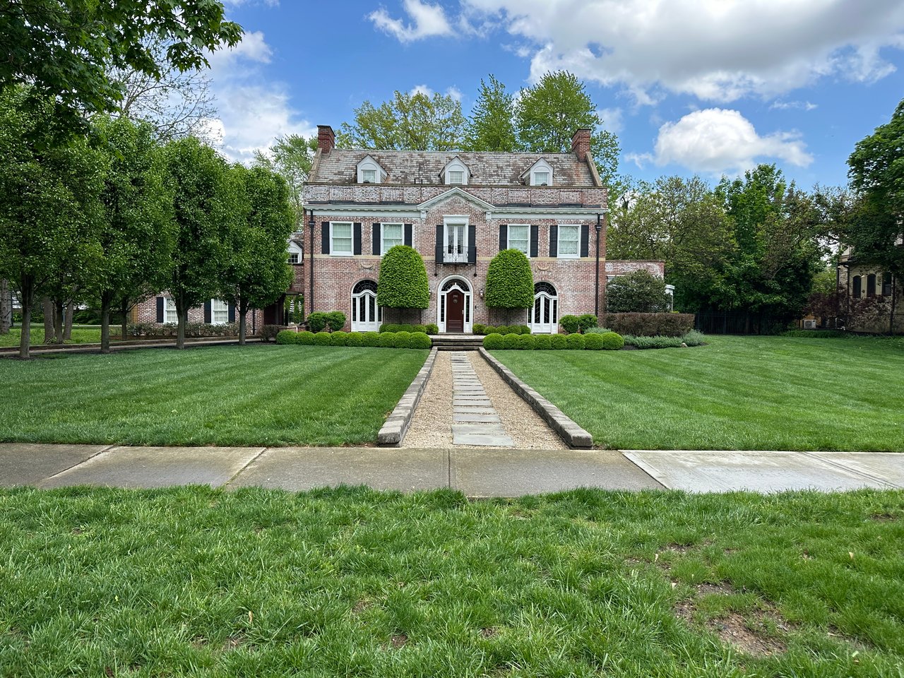 Brick two-story home with a slate roof, dormers, shaped shrubs, and a stone walkway across the lawn.