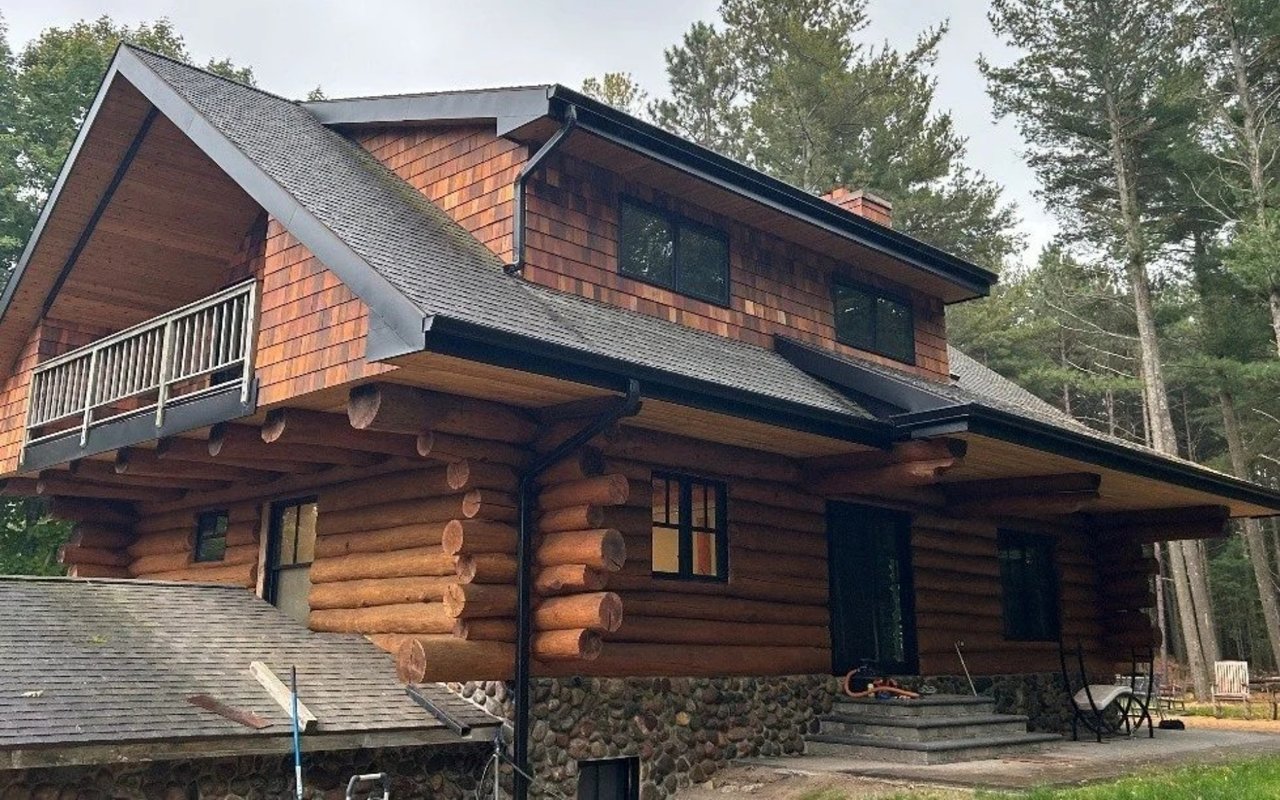 Log home with a stone foundation, shingled upper story, and balcony among tall trees.