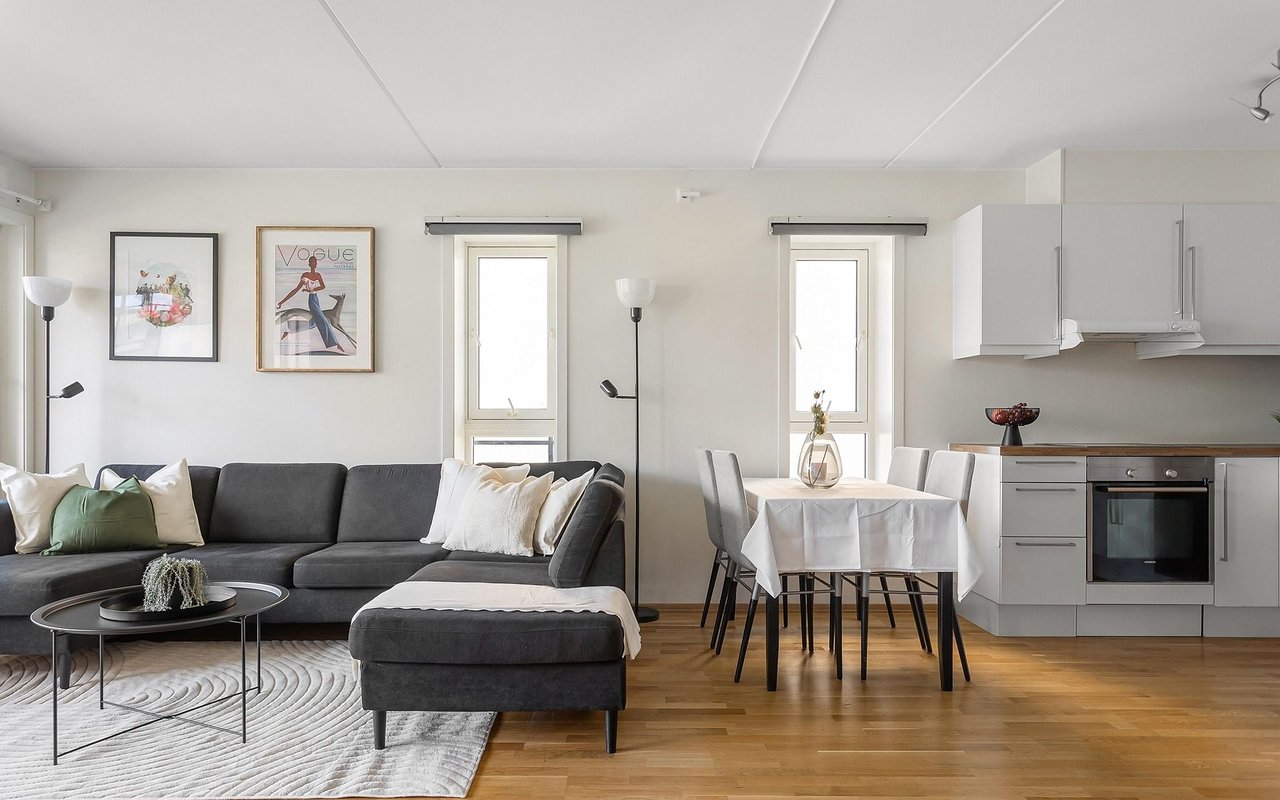 Open-plan living and dining area with a gray sectional, white kitchen, and warm wood floors.