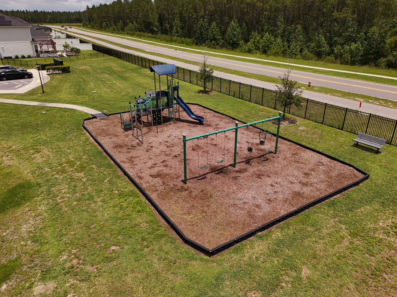 Neighborhood playground with a blue-roofed play structure, slide, and swings inside a fenced wood-chip area.