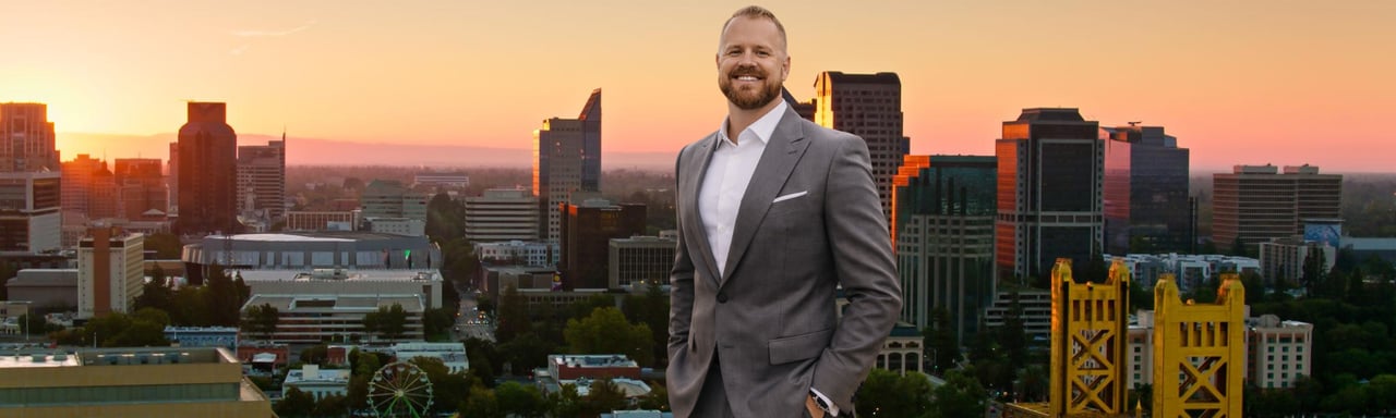 Smiling real estate agent in a gray suit posed before the Sacramento skyline at sunset.