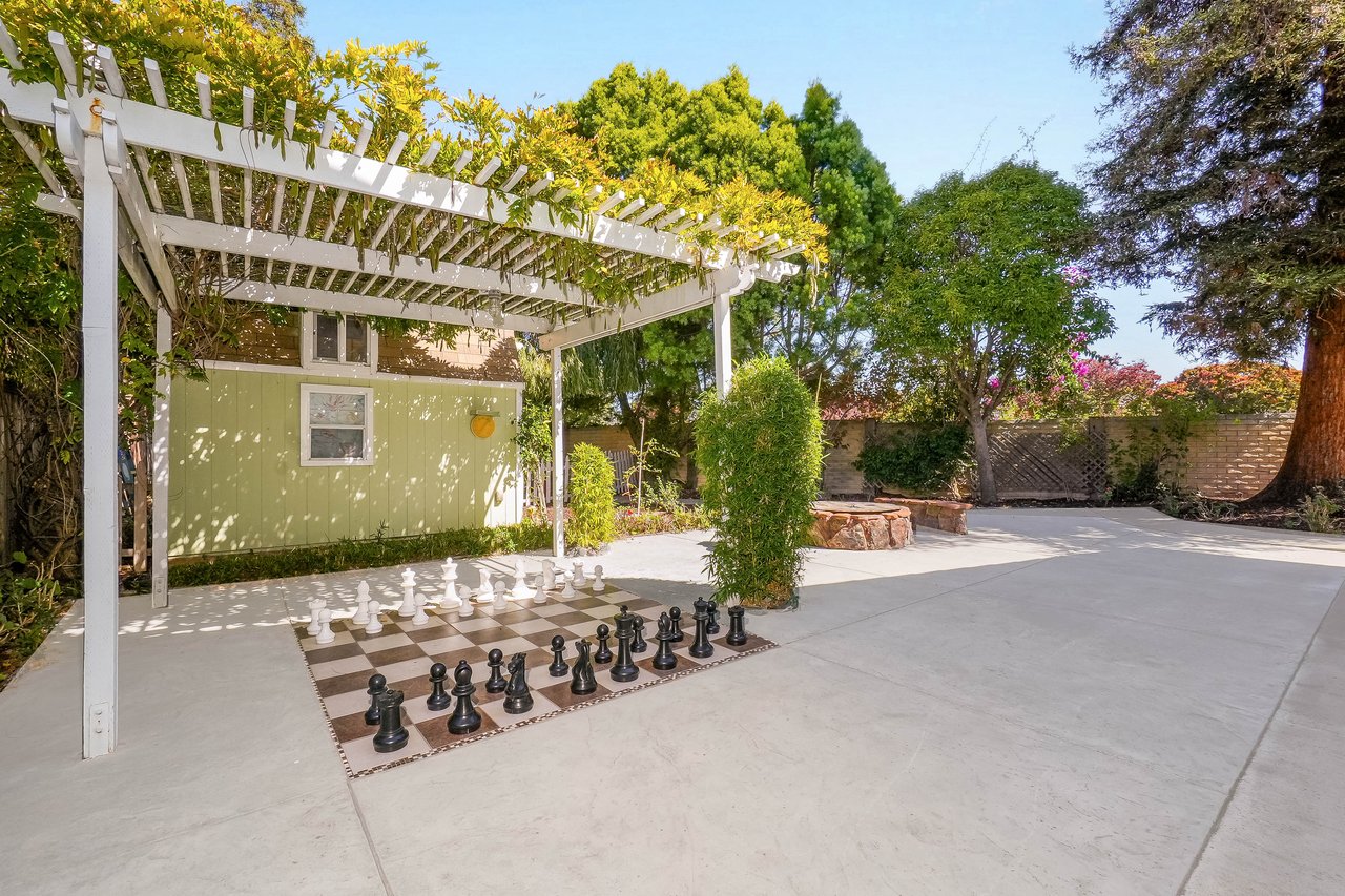 Backyard patio with a vine-covered pergola, oversized chess set, and circular stone fire pit.