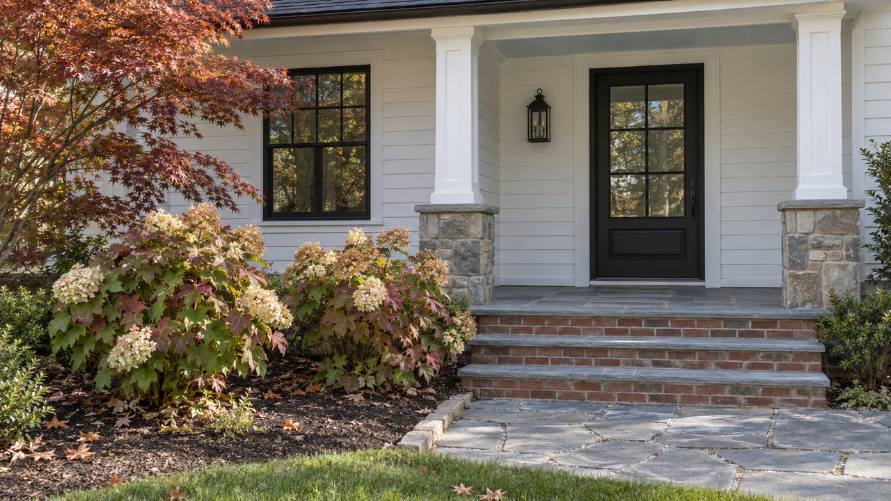 White clapboard home with a black glazed entry, bluestone steps, and autumn oakleaf hydrangeas beside a garden path.
