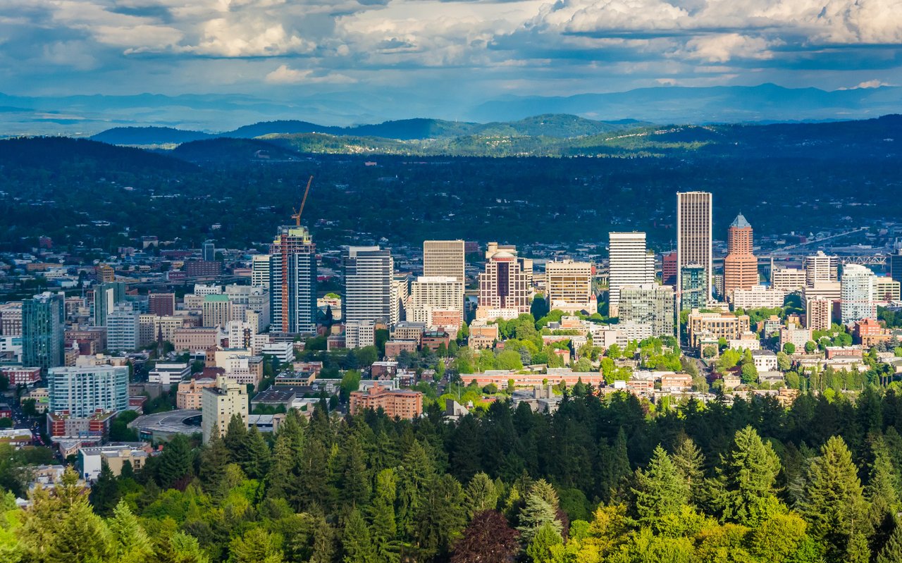 Portland skyline framed by green treetops, forested hills, and a cloudy blue sky.