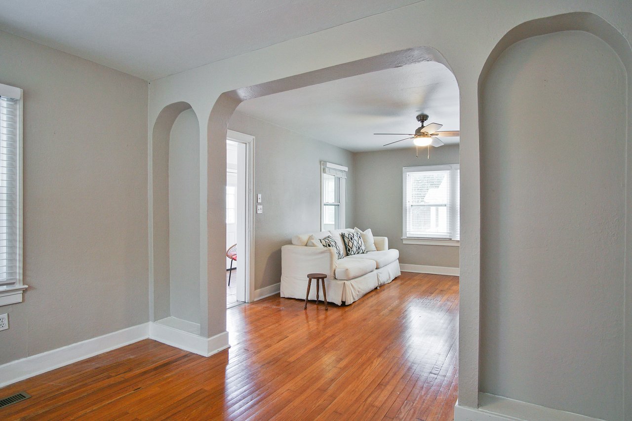 Arched doorway frames a cozy living room with hardwood floors, a white sofa, decorative pillows, and a ceiling fan. Large windows bring in natural light.