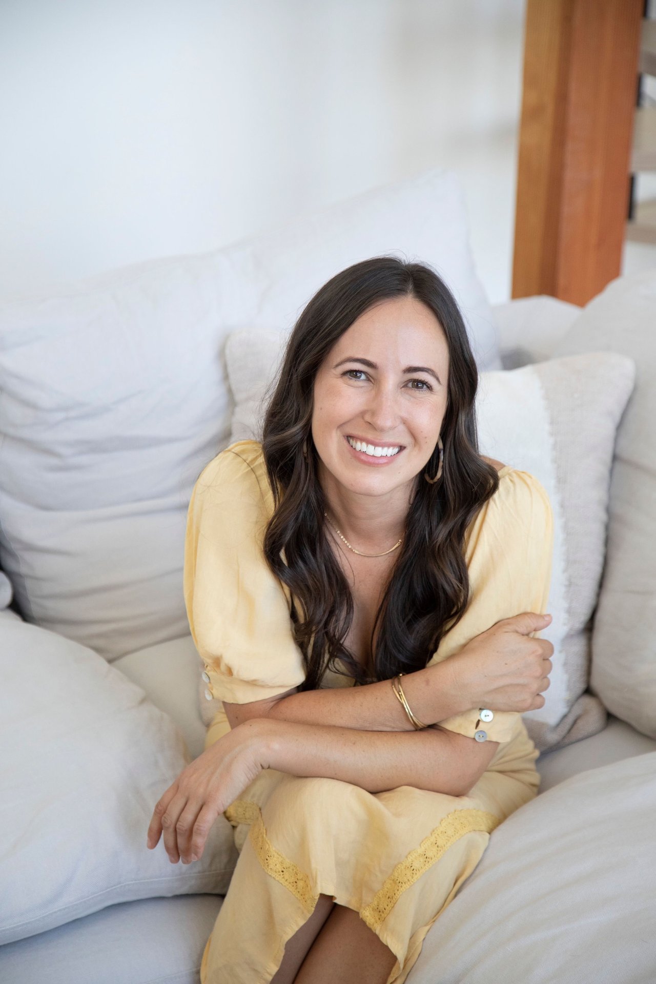 Veronica Lewis smiles while seated on a white sofa in a pale yellow dress.
