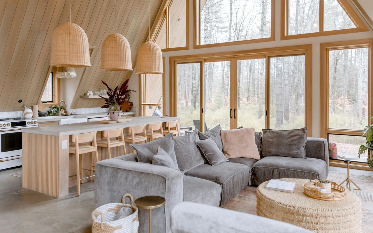 Open-plan living room and kitchen with a vaulted wood ceiling, gray sectional, and large windows overlooking trees.