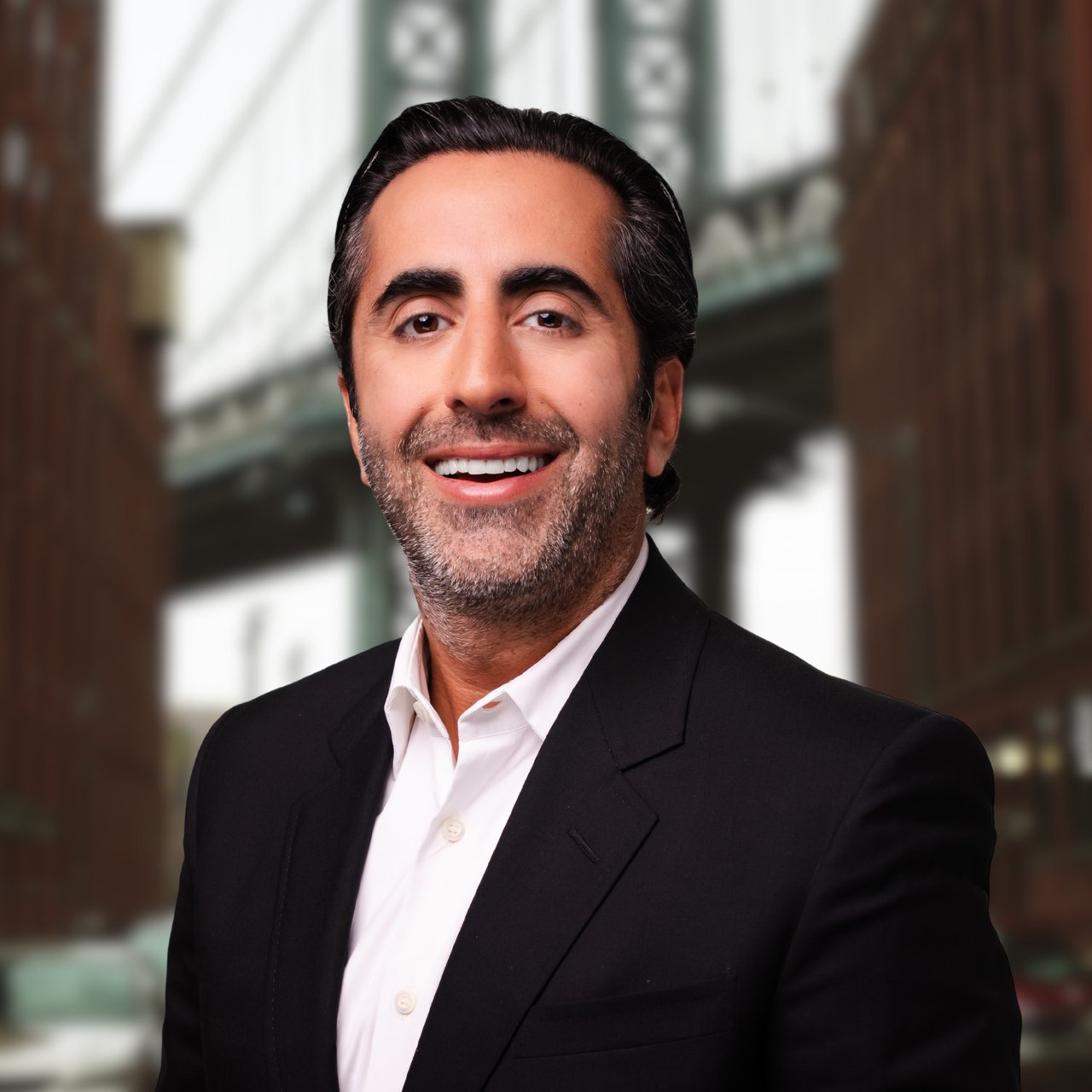 Khashy Eyn in a black suit jacket and white shirt, smiling against a blurred architectural background.