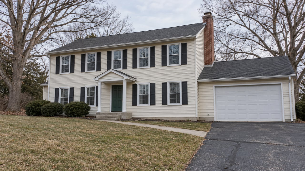 Cream-sided two-story home with black shutters, an attached garage and a sloping winter lawn beneath bare oak trees.