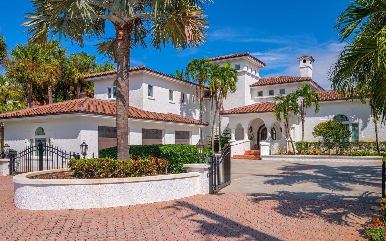 White stucco residence with red tile roofs, arched entryway, palm trees, and a gated driveway.