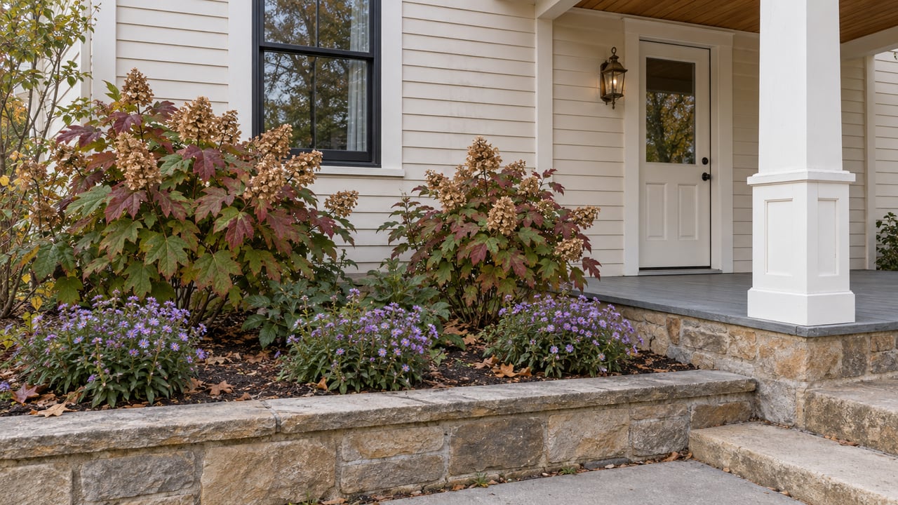 Cream clapboard home with a covered entry, weathered sandstone garden wall, oakleaf hydrangeas and violet asters in soft autumn daylight.