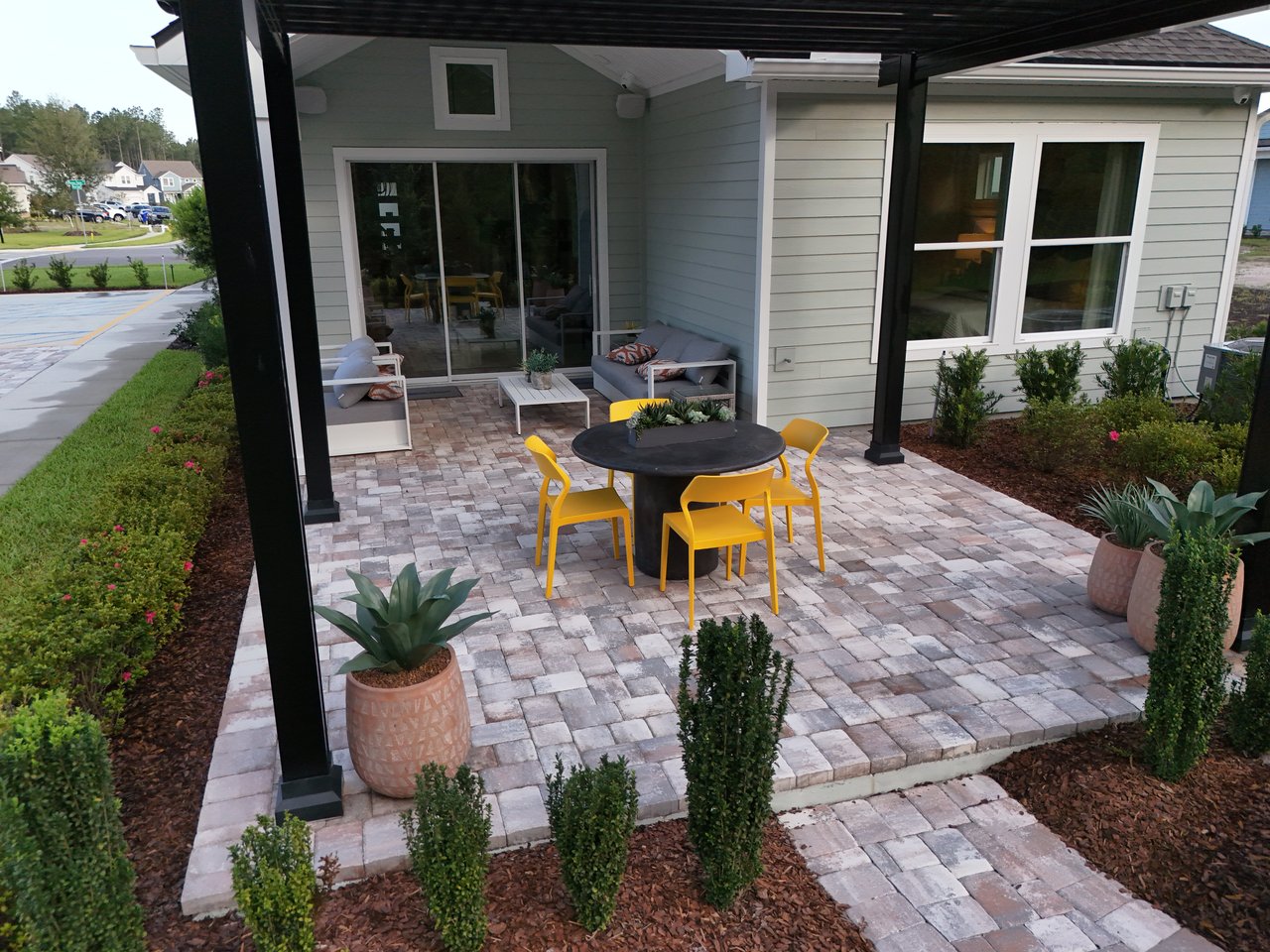 Covered paver patio with yellow dining chairs, a round table, and lounge seating beside the house.
