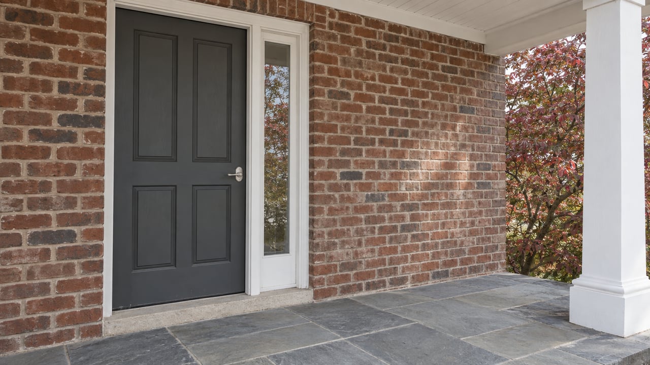 Brick home entrance with a charcoal paneled door, white porch column, gray-blue stone paving, and autumn dogwood leaves.