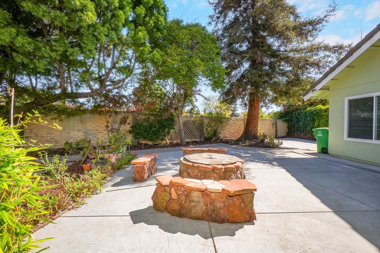Backyard patio with a circular stone fire pit, matching benches, mature trees, and garden beds.