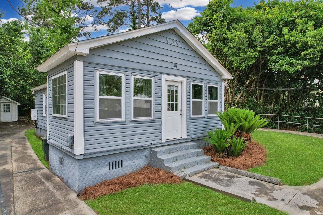 A cozy, light blue house with white trim sits amidst lush greenery. Steps lead to a white door, framed by windows. A tidy lawn and shrubs add charm.