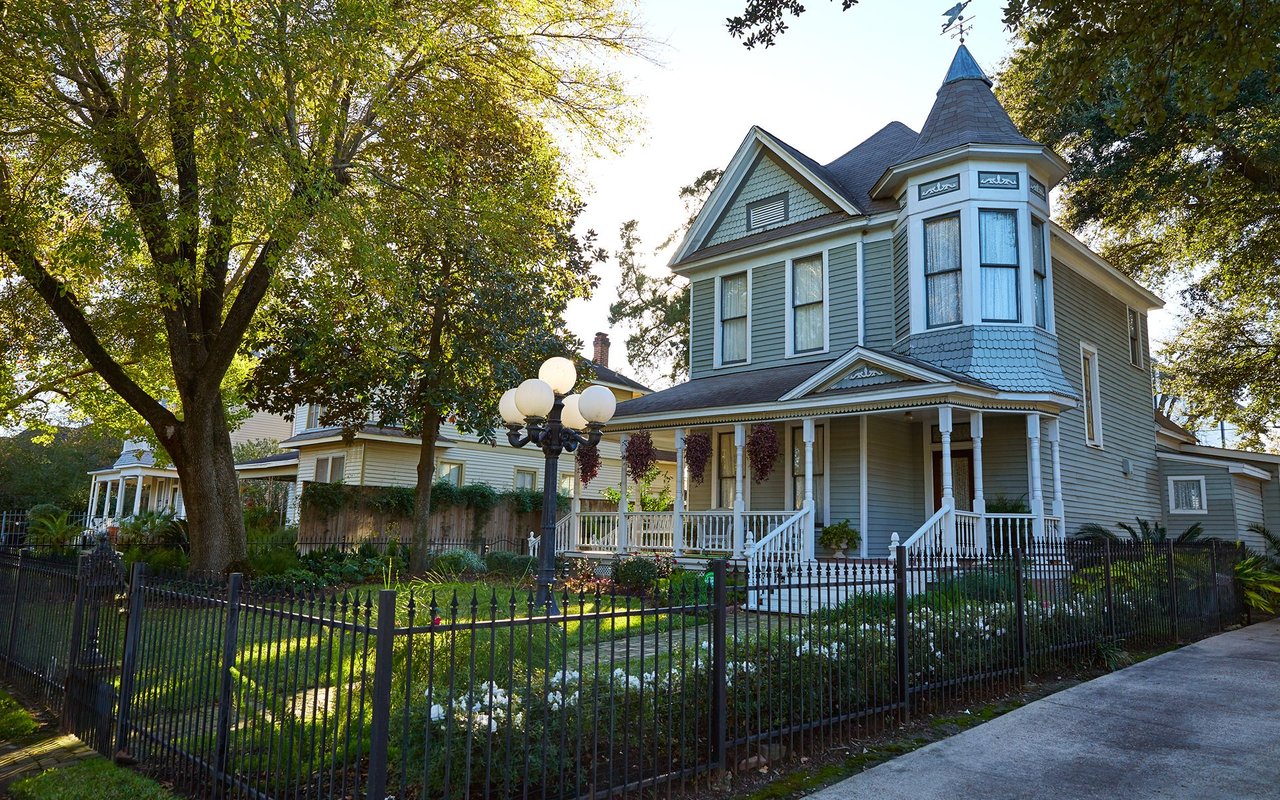 Blue-gray Victorian house with a turret, wraparound porch, and fenced garden beneath mature trees.