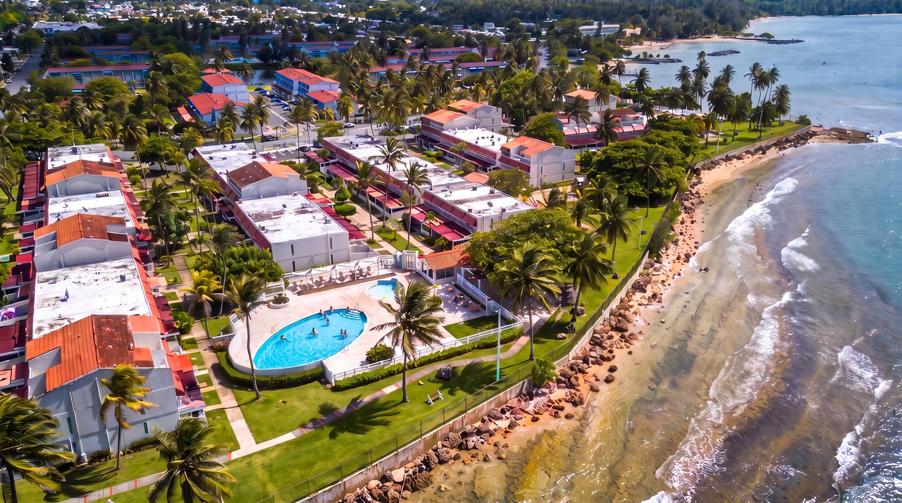Coastal residential complex with a curved swimming pool, palm trees, and rocky shoreline.