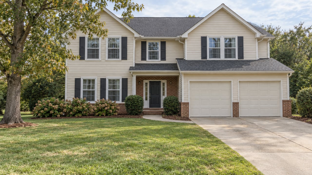 Ivory-sided two-story home with brick accents, dark shutters, a broad front lawn and a mature oak under bright cloud-filtered daylight.