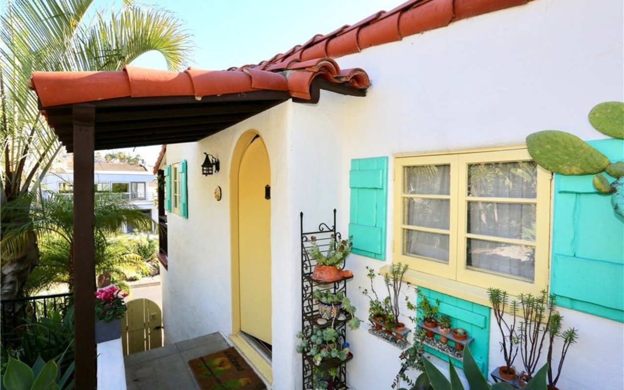 Home entry with a yellow arched door, turquoise shutters, and potted plants beneath a red tile roof.