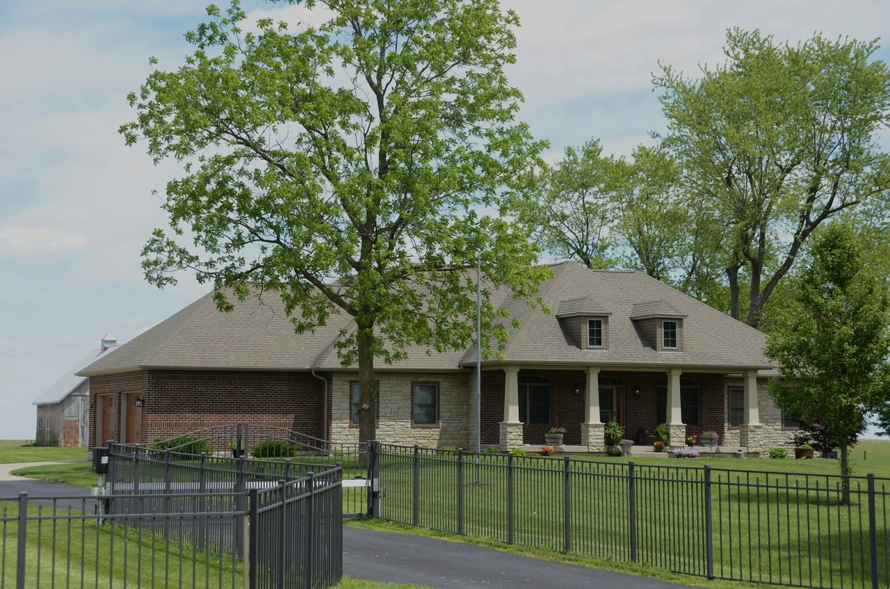 Brick and stone home with a covered porch, two dormers, mature trees, and fenced lawn.