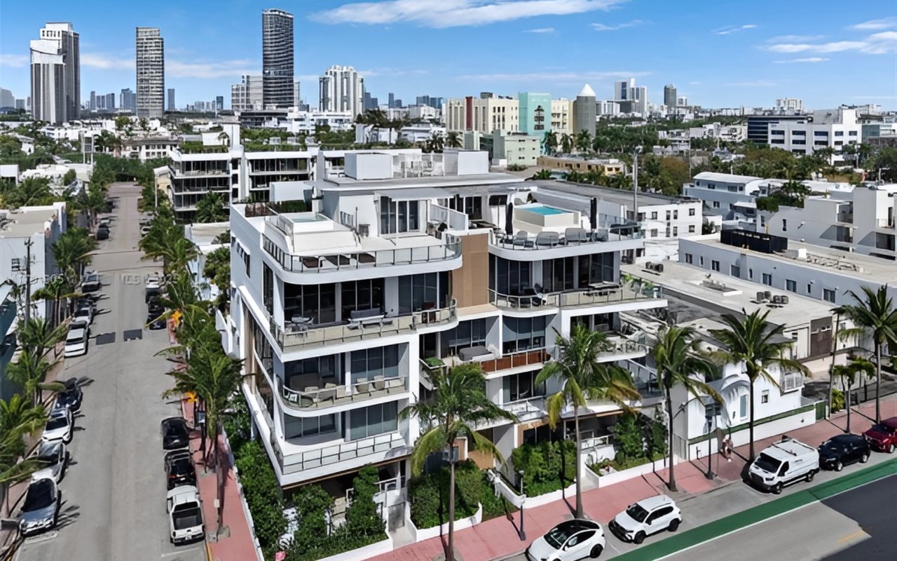 Aerial view of a white residential building with balconies and a rooftop pool, with the city skyline beyond.