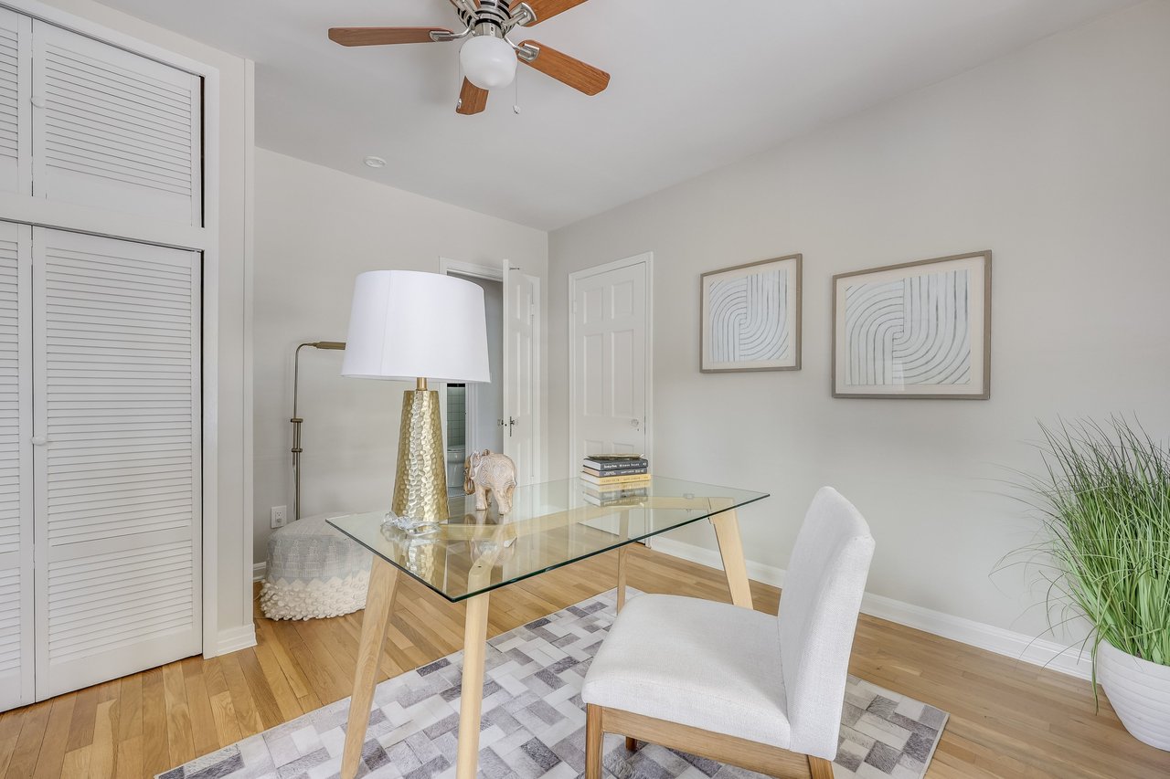 Staged home office with a glass desk, white chair, hardwood floors, and louvered closet doors.