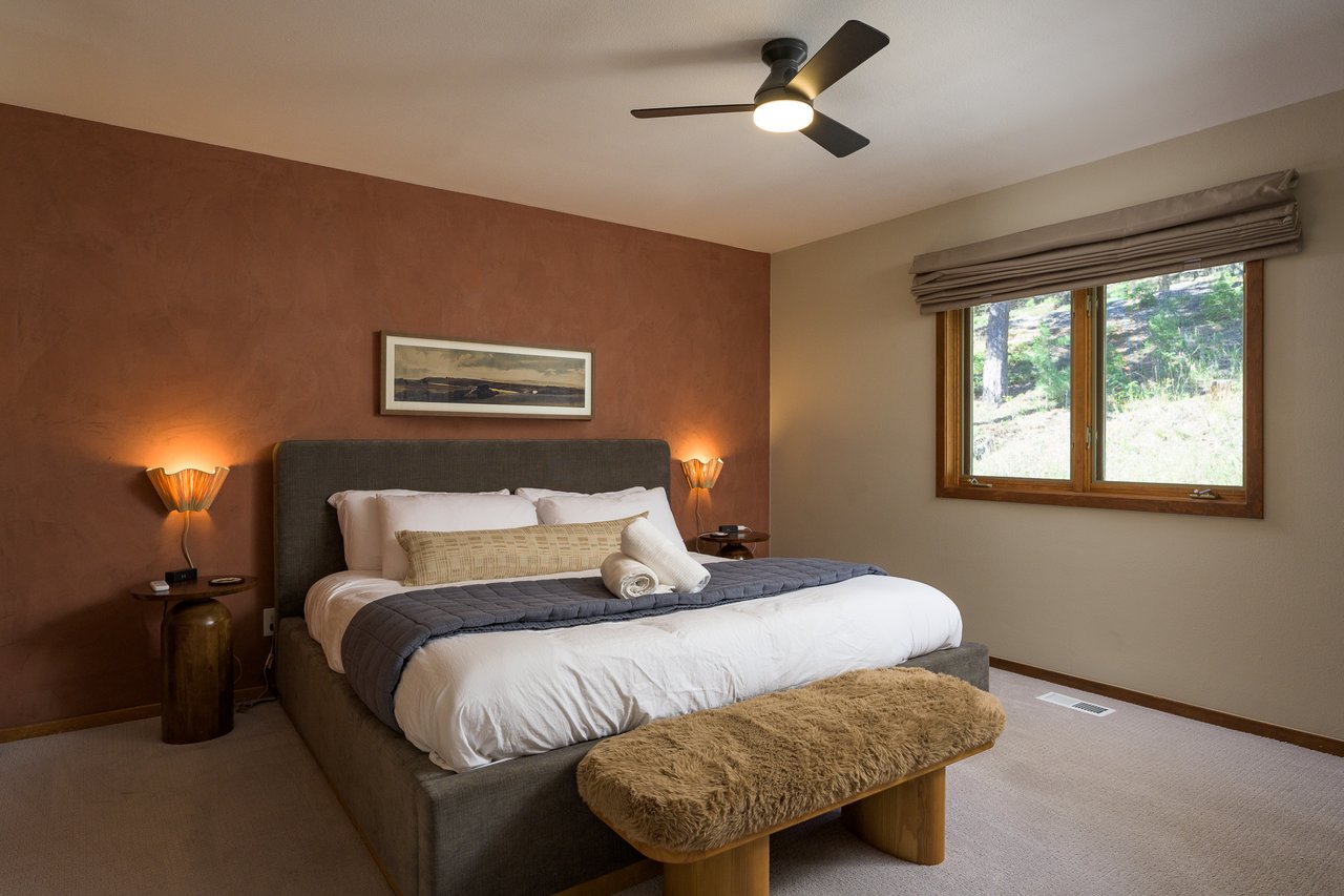 Bedroom with a large bed against a rust-colored accent wall and a wood-framed window overlooking trees.