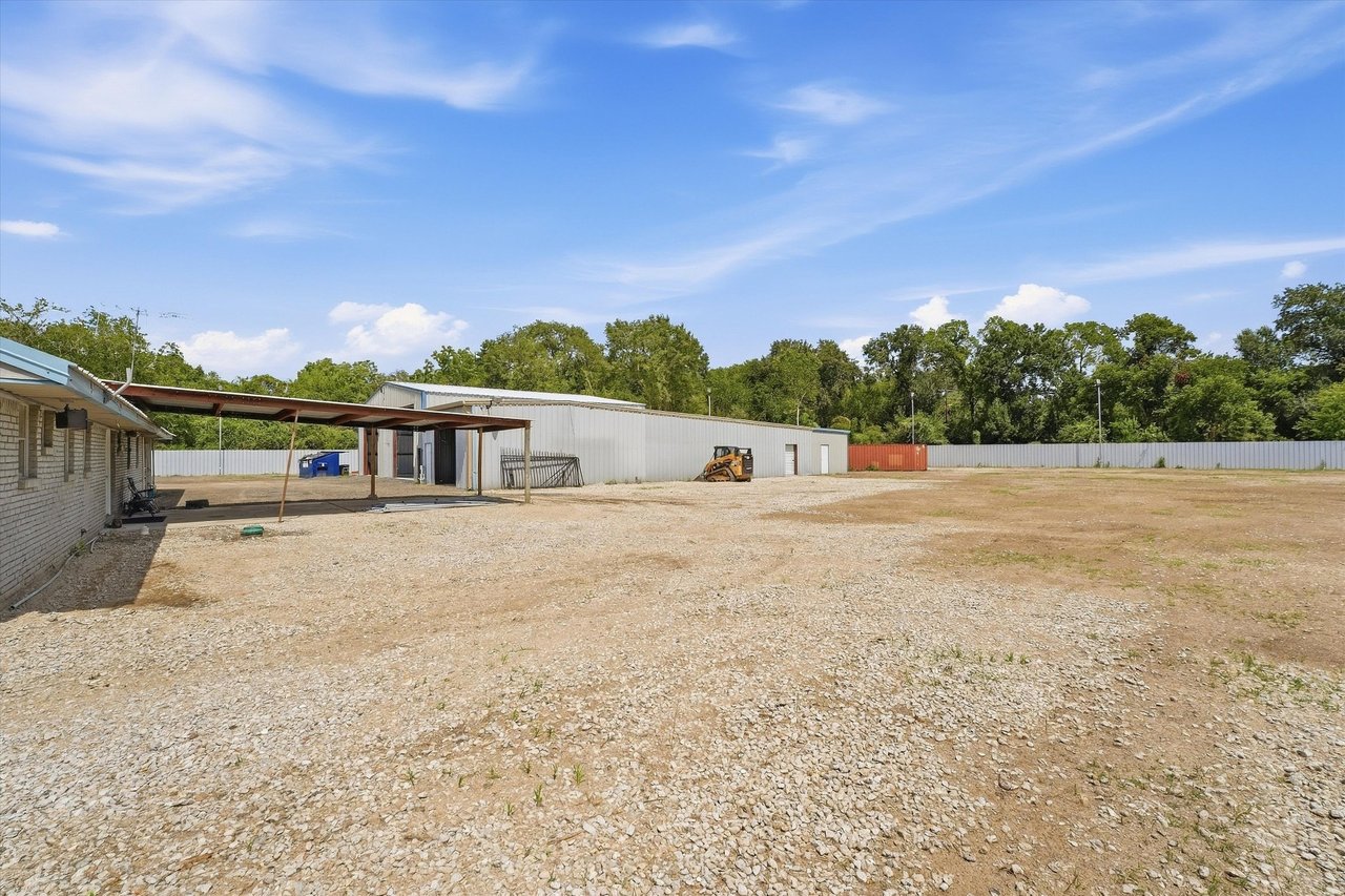 Fenced gravel lot beside a long metal warehouse and covered work area.