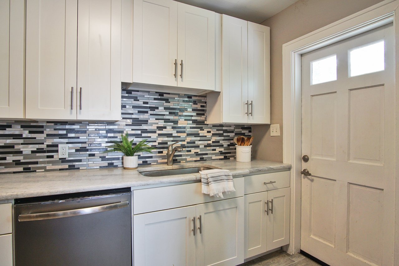 Modern kitchen with white cabinets, gray countertops, and stainless steel dishwasher. A mosaic tile backsplash adds contrast. A potted plant and utensils provide a cozy touch.
