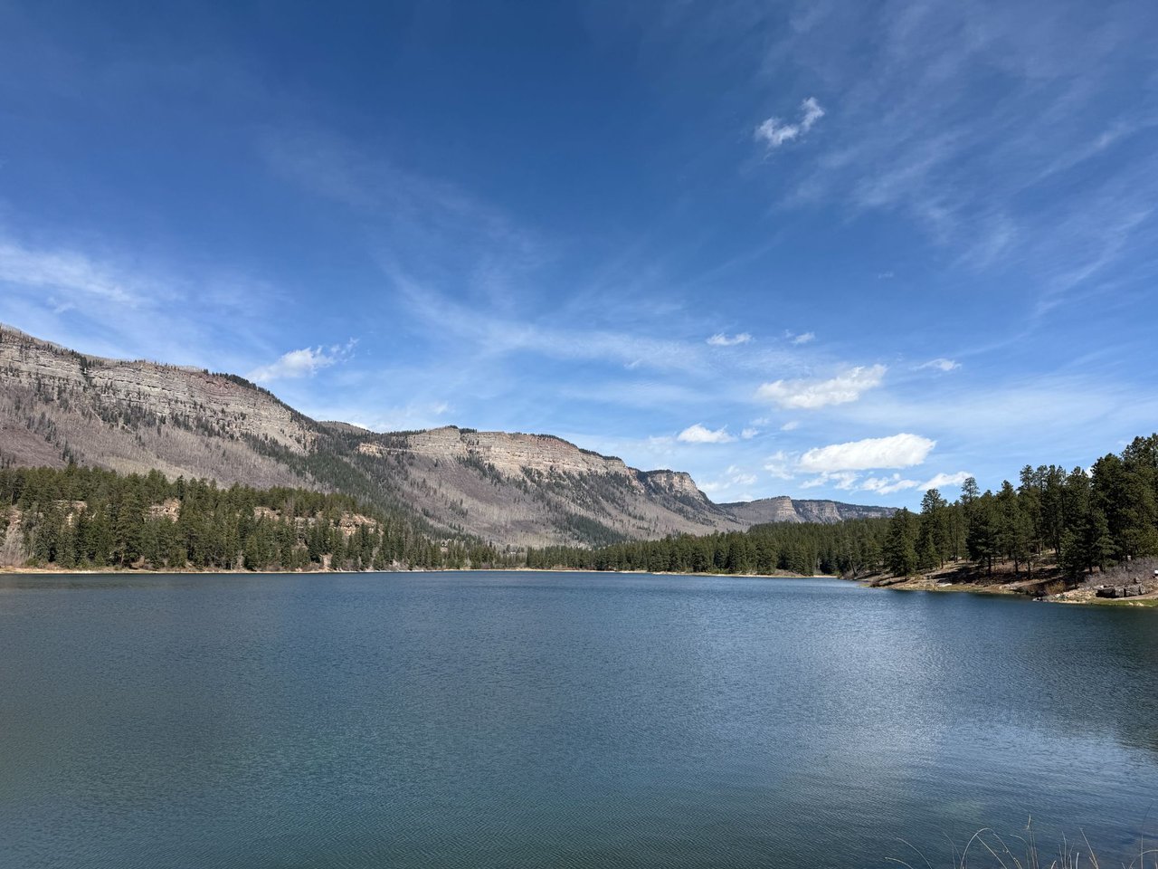 Mountain lake bordered by pine trees and rocky ridges beneath a broad blue sky.