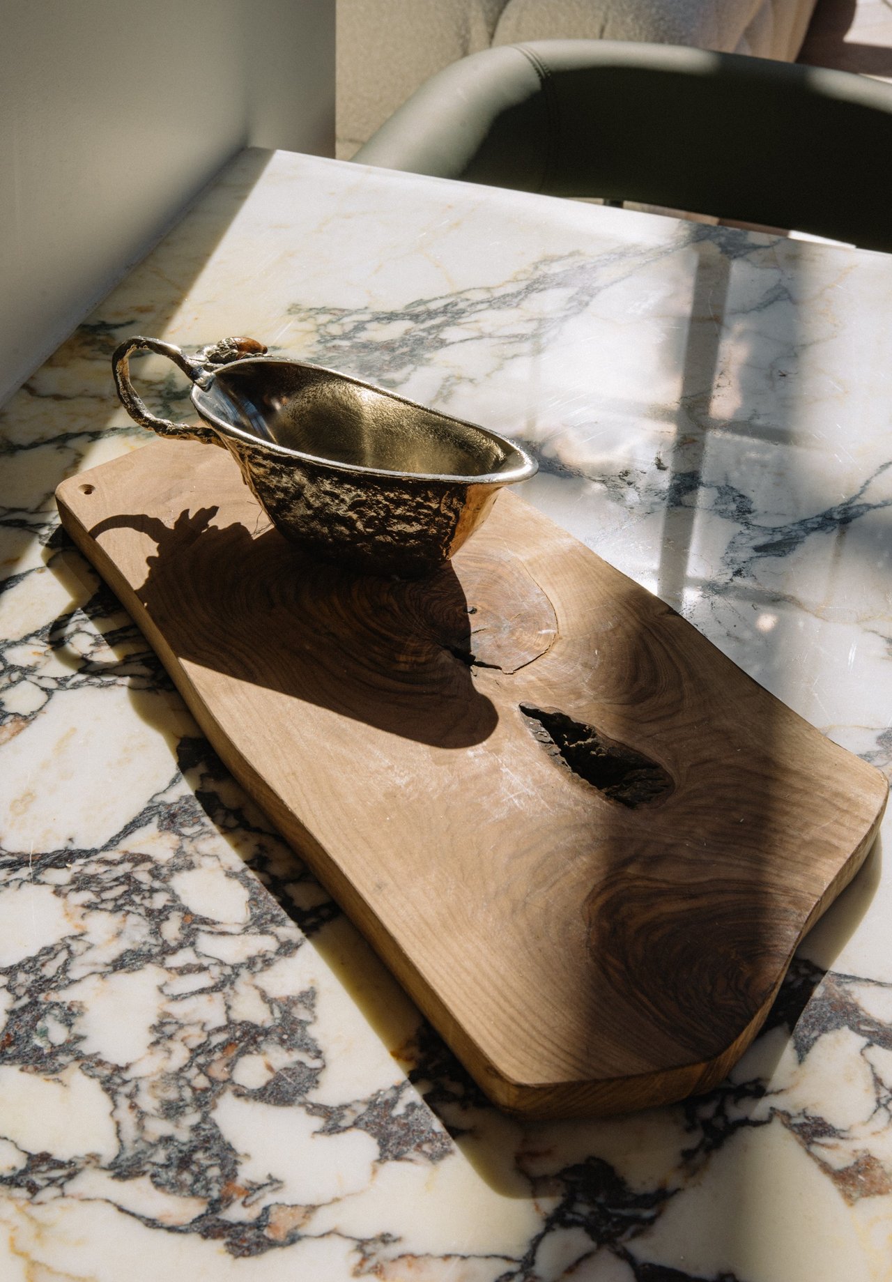 Metal vessel on a live-edge wooden board atop a veined marble counter in sunlight.